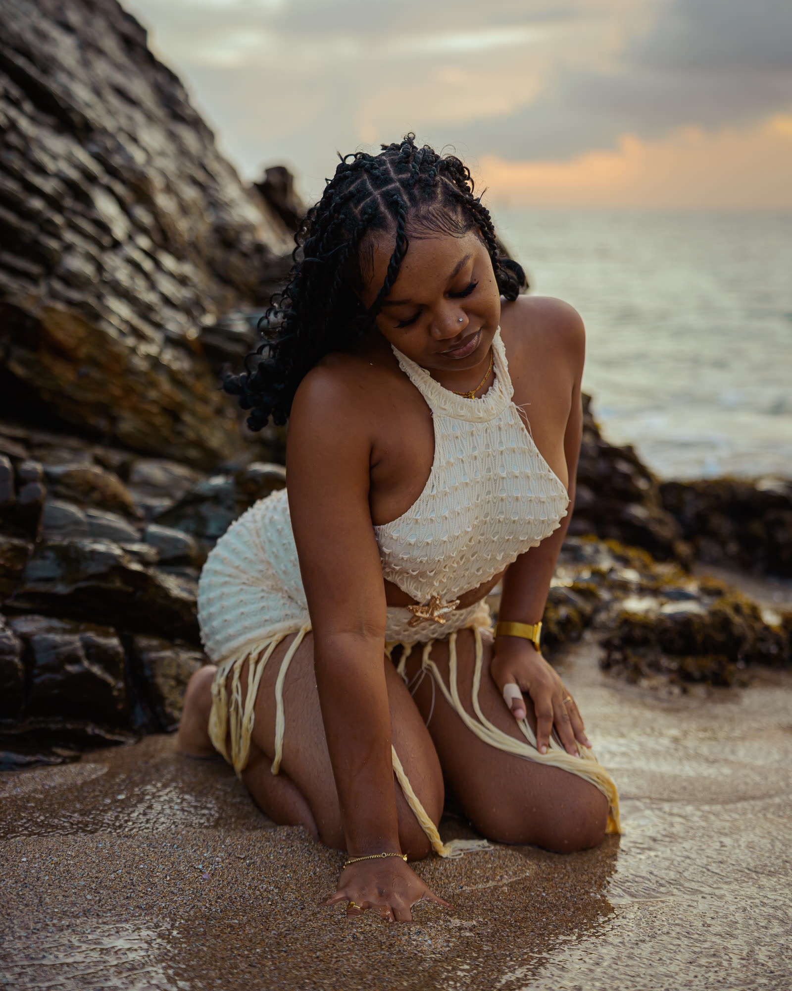 Loana à genoux sur le sable au pied des rochers, au coucher du soleil